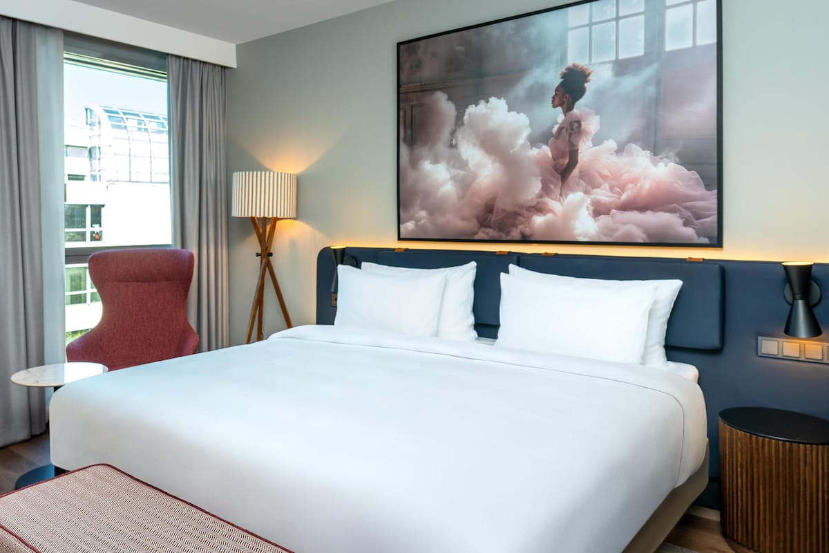 A hotel room with a large bed, a red chair, a small table, and a large painting on the wall.