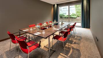A conference room with a long table, red chairs, and a view of greenery outside.