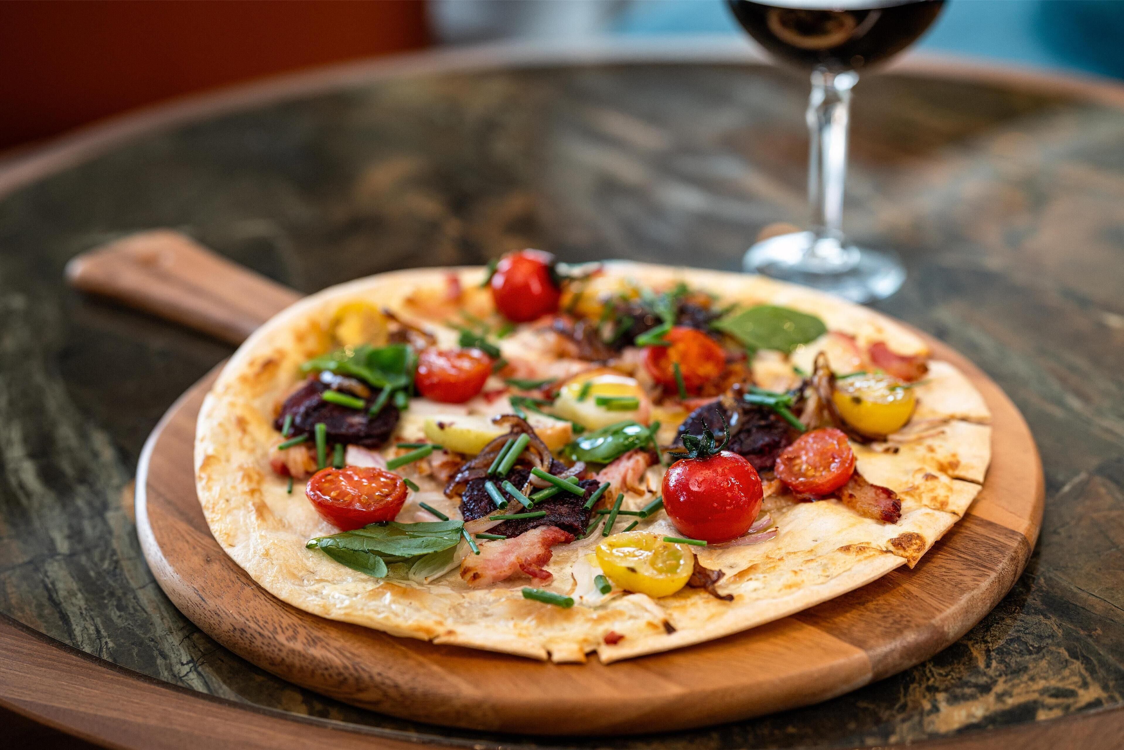 A pizza on a wooden board with various toppings, a glass of red wine, and a rustic table surface.