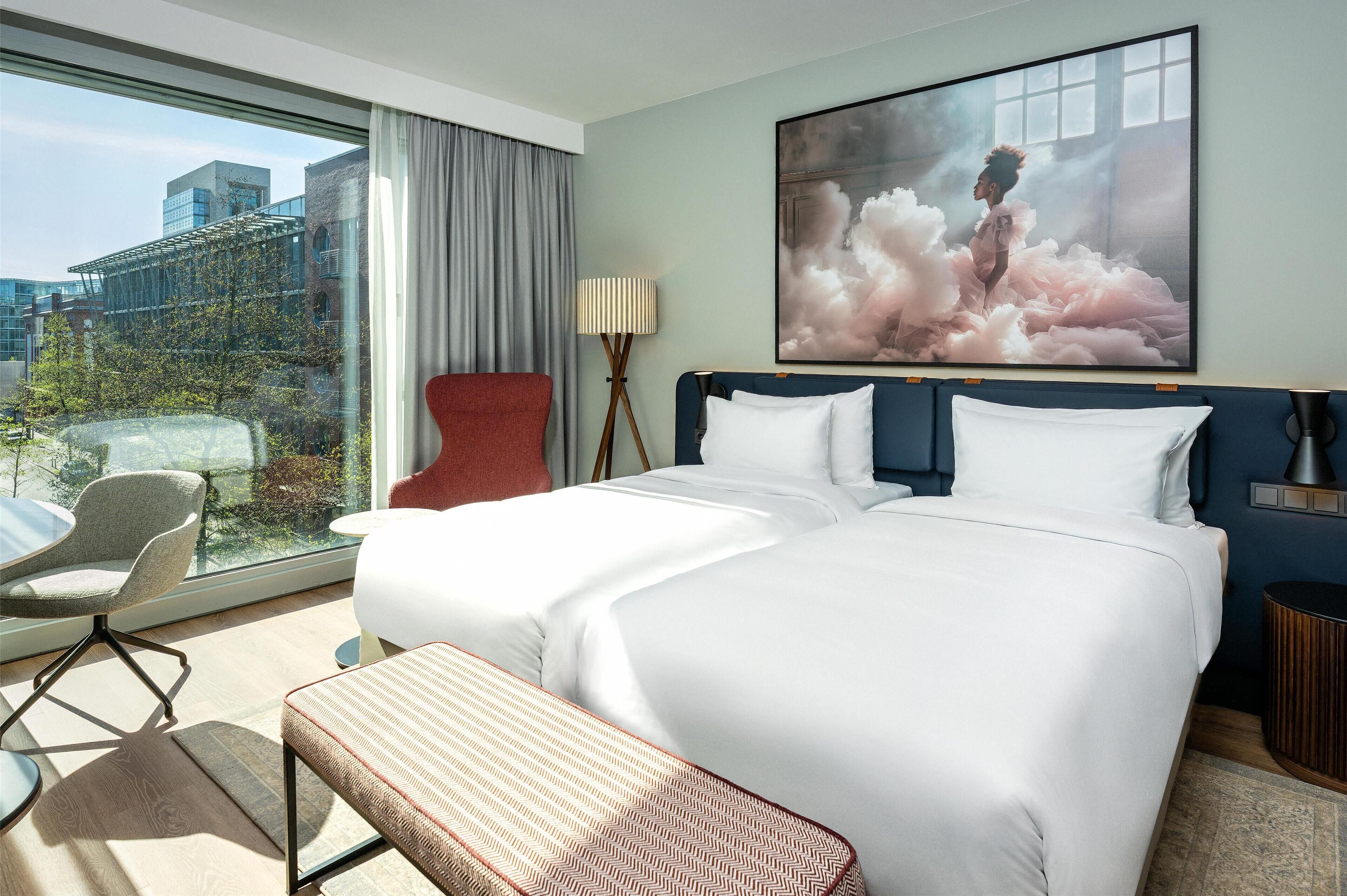 Ein Hotelzimmer mit einem großen Bett, einem Schreibtisch, einem Stuhl, einer Bank, einer Lampe und einem großen Fenster mit Blick auf Gebäude und Bäume.