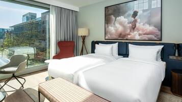 Ein Hotelzimmer mit einem großen Bett, einem Schreibtisch, einem Stuhl, einer Bank, einer Lampe und einem großen Fenster mit Blick auf Gebäude und Bäume.