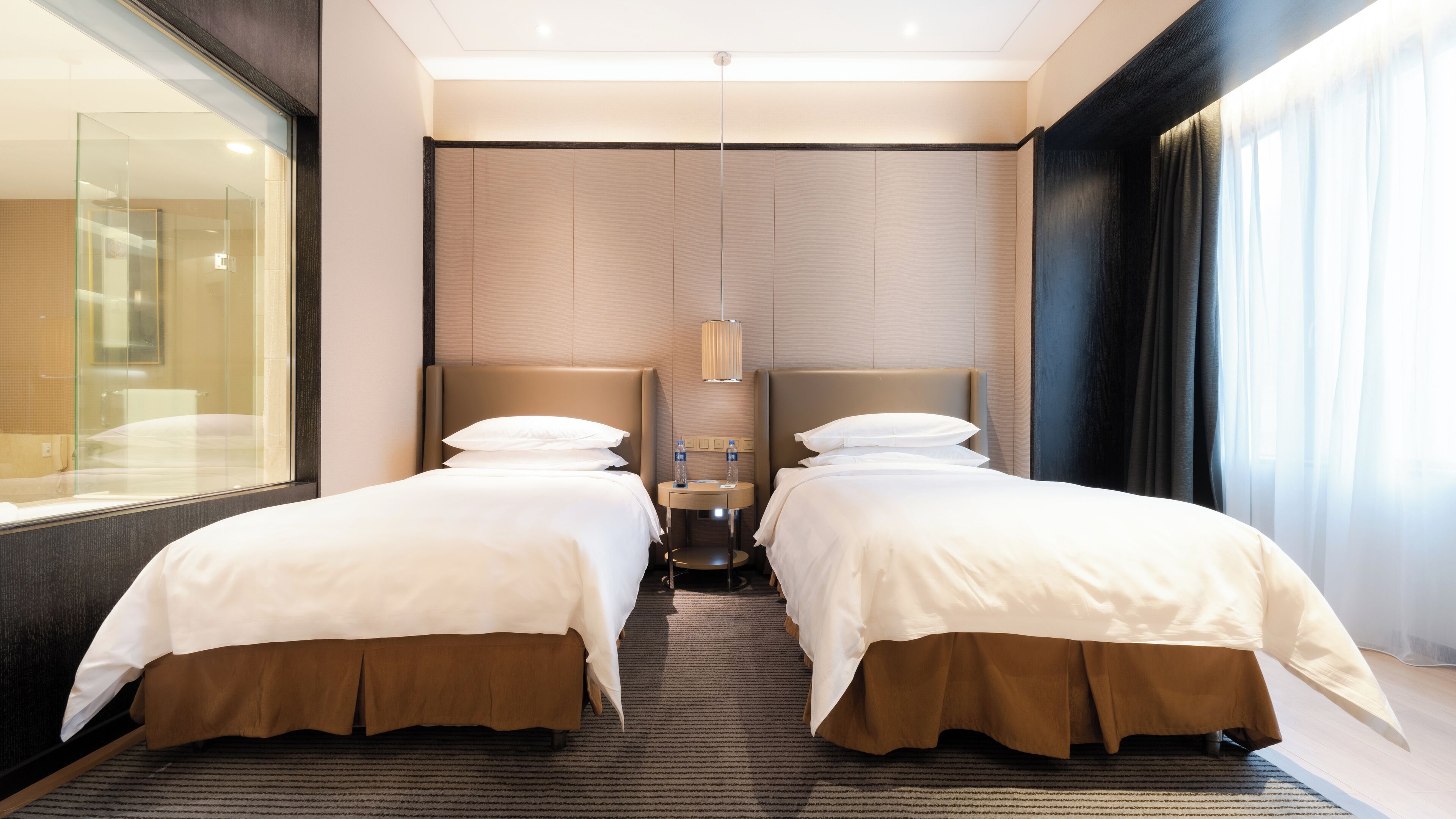 Une chambre d’hôtel avec deux lits, une petite table sur laquelle sont posées deux bouteilles, une lampe fixée au mur et une grande fenêtre avec des rideaux.