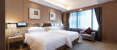 Habitación de hotel con dos camas, un escritorio con lámpara, una silla y un gran ventanal con cortinas.