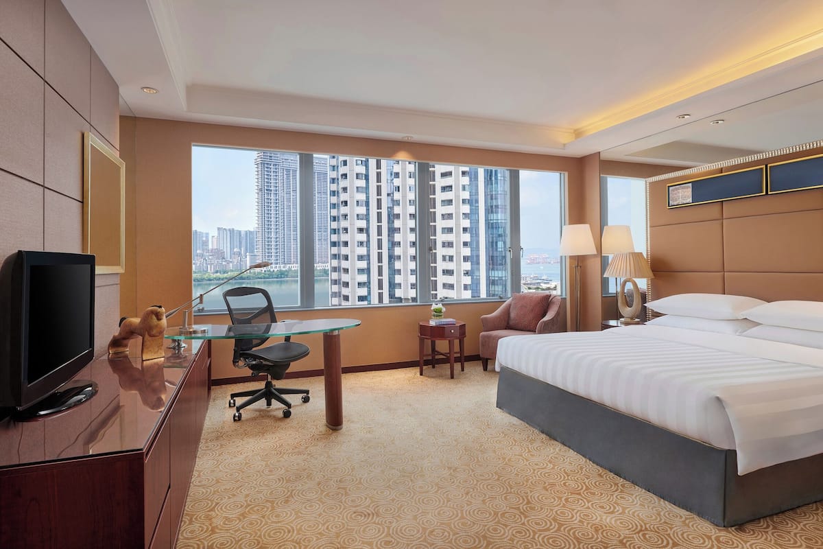 Ein Hotelzimmer mit einem großen Bett, einem Schreibtisch mit Computer, einem Fernseher und Blick auf hohe Gebäude.