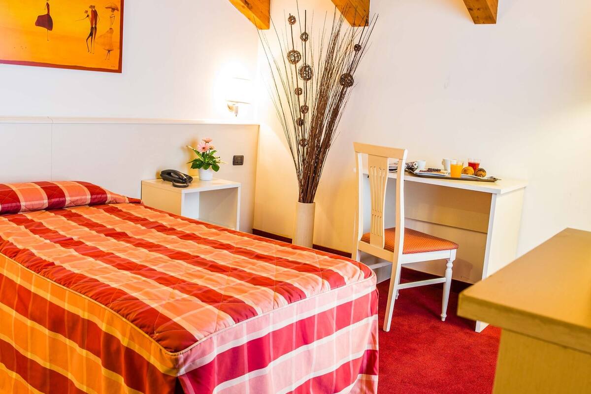 A hotel room with a bed, a desk, a chair, a vase with dried plants, and a painting on the wall.