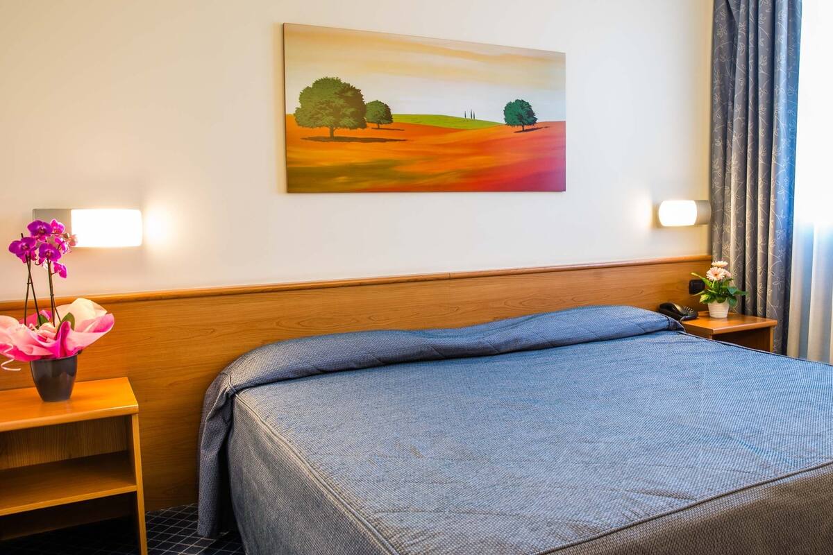 A hotel room with a bed, a nightstand with a phone, a vase with flowers, and a painting on the wall.