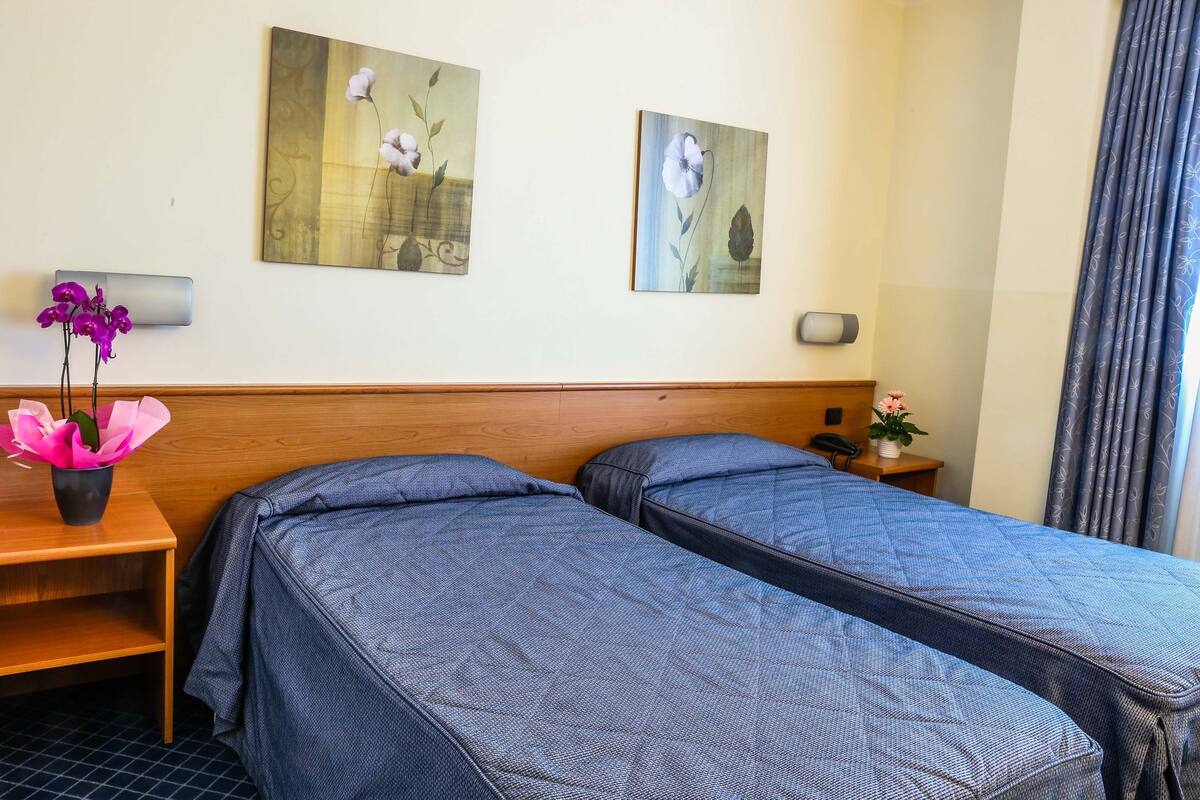 A hotel room with two beds, a wooden headboard, a nightstand with a phone, a vase with flowers, and two wall-mounted paintings.