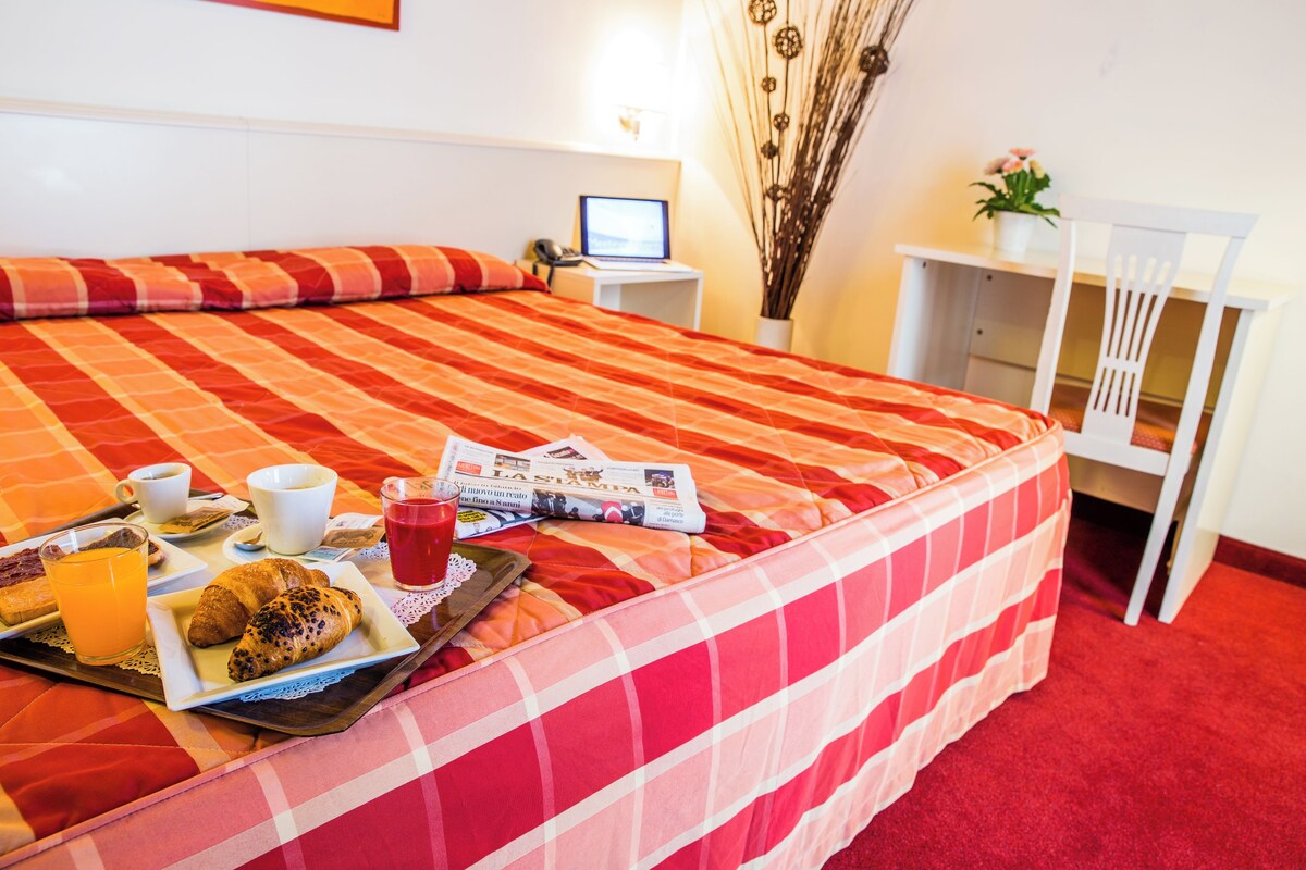 A hotel room with a large bed, a bedside table with a laptop, a tray with breakfast items, and a chair with a small table.