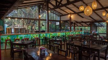 Un restaurante con zona de bar, mesas y sillas de madera, y grandes ventanales que ofrecen vistas al exterior.