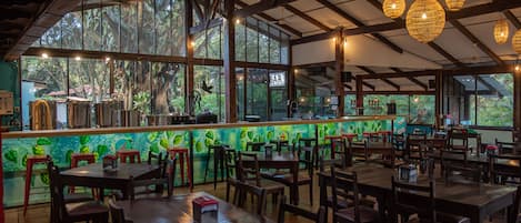 A restaurant with a bar area, wooden tables and chairs, and large windows offering a view of the outdoors.