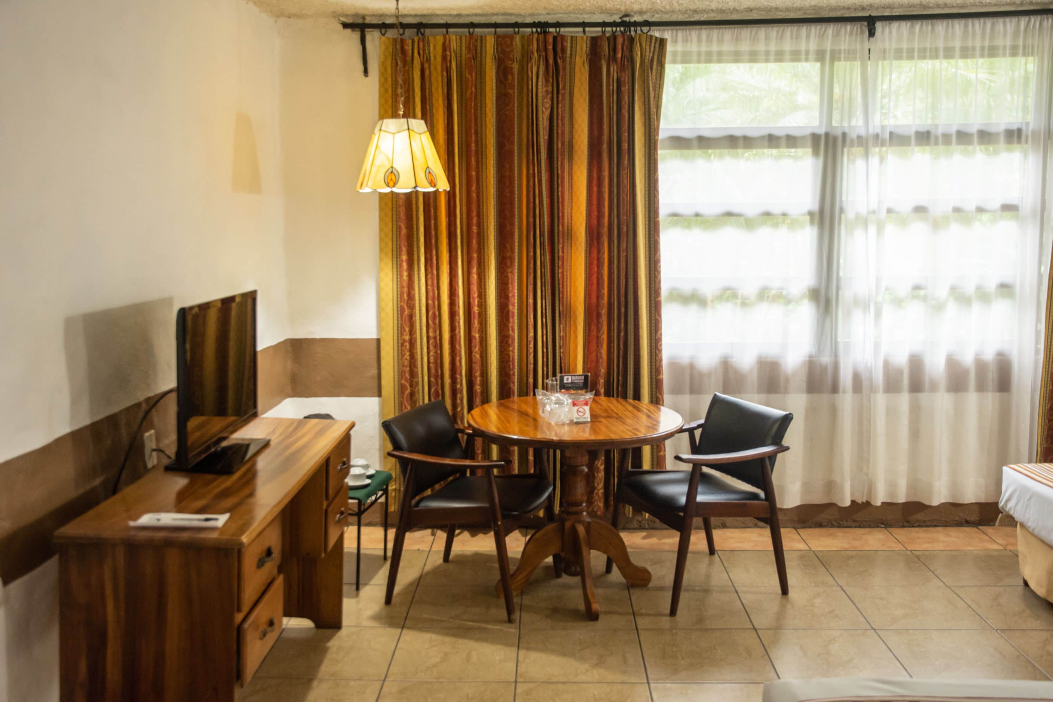Une pièce comprenant une table à manger en bois, quatre chaises, un téléviseur posé sur un meuble en bois et une fenêtre avec des rideaux.