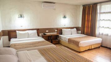 A hotel room with two beds, a bedside table with a lamp, a wall-mounted air conditioner, and a window with curtains.