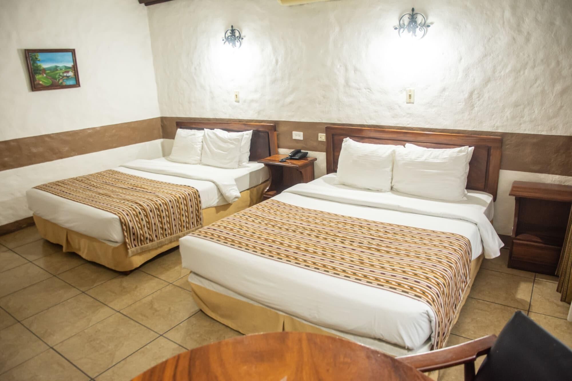 A hotel room with two beds, a wooden nightstand with a phone, a small table, and a wall-mounted light fixture.