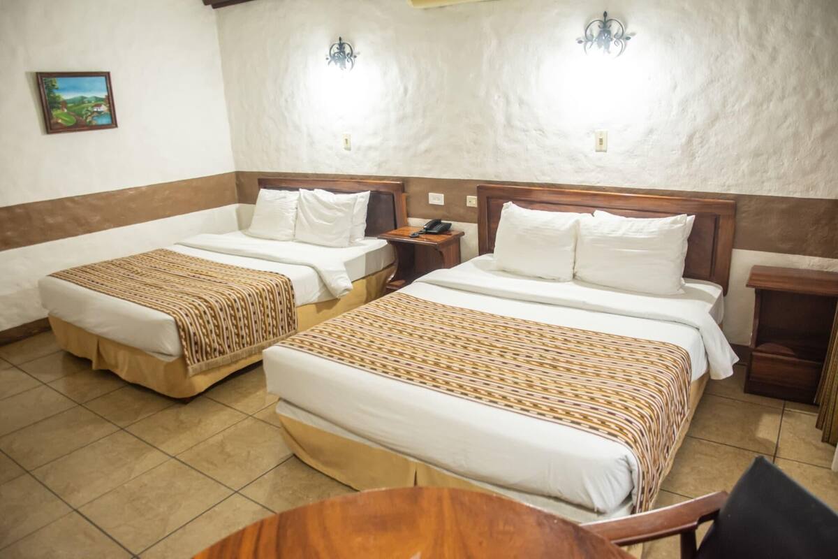 A hotel room with two beds, a wooden nightstand with a phone, a small table, and a wall-mounted light fixture.
