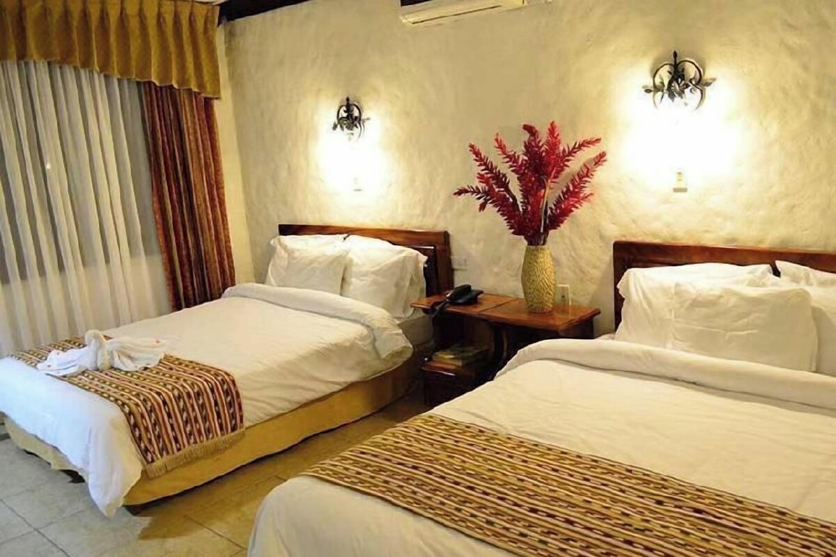 A hotel room with two beds, a nightstand, a vase with red flowers, and a wall-mounted light fixture.