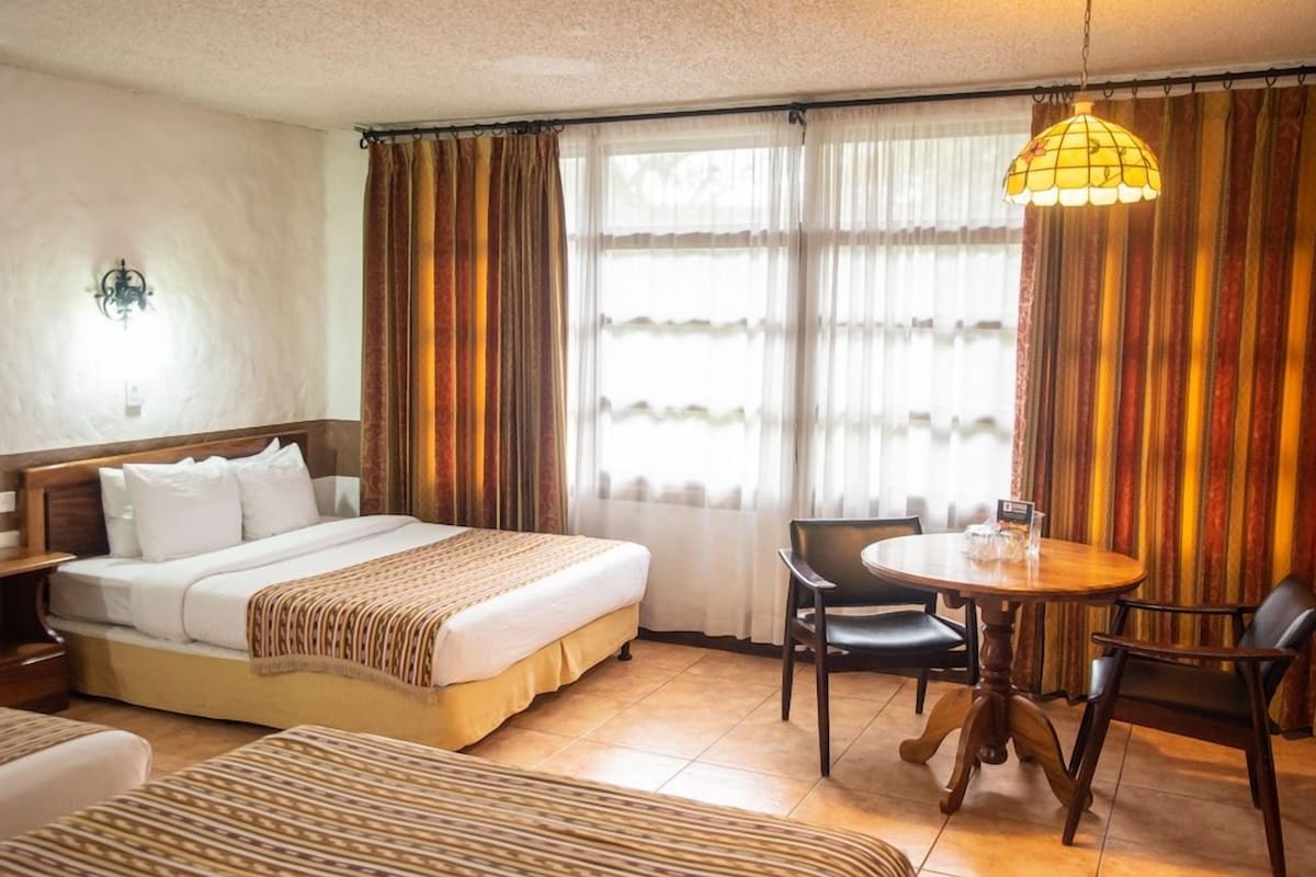 A hotel room with a bed, bedside table, lamp, curtains, and a dining table with chairs.