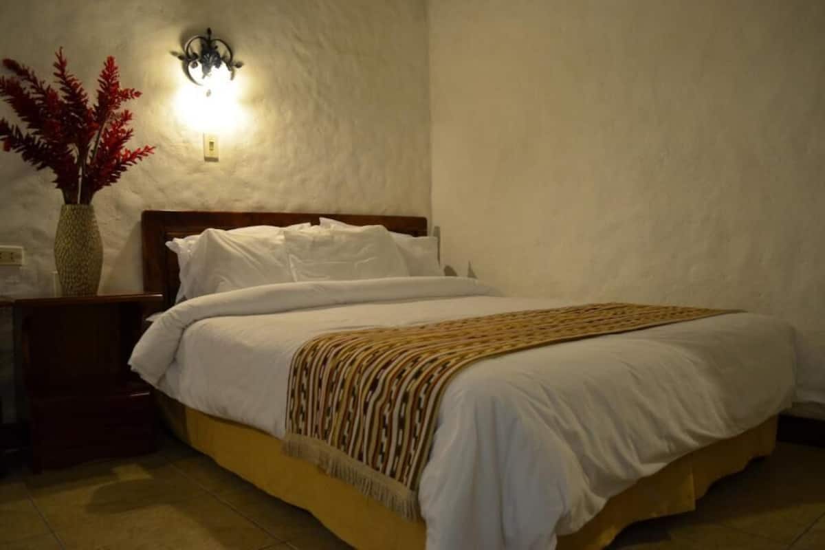 A bedroom with a bed, a nightstand, a wall-mounted light, and a vase with red foliage.