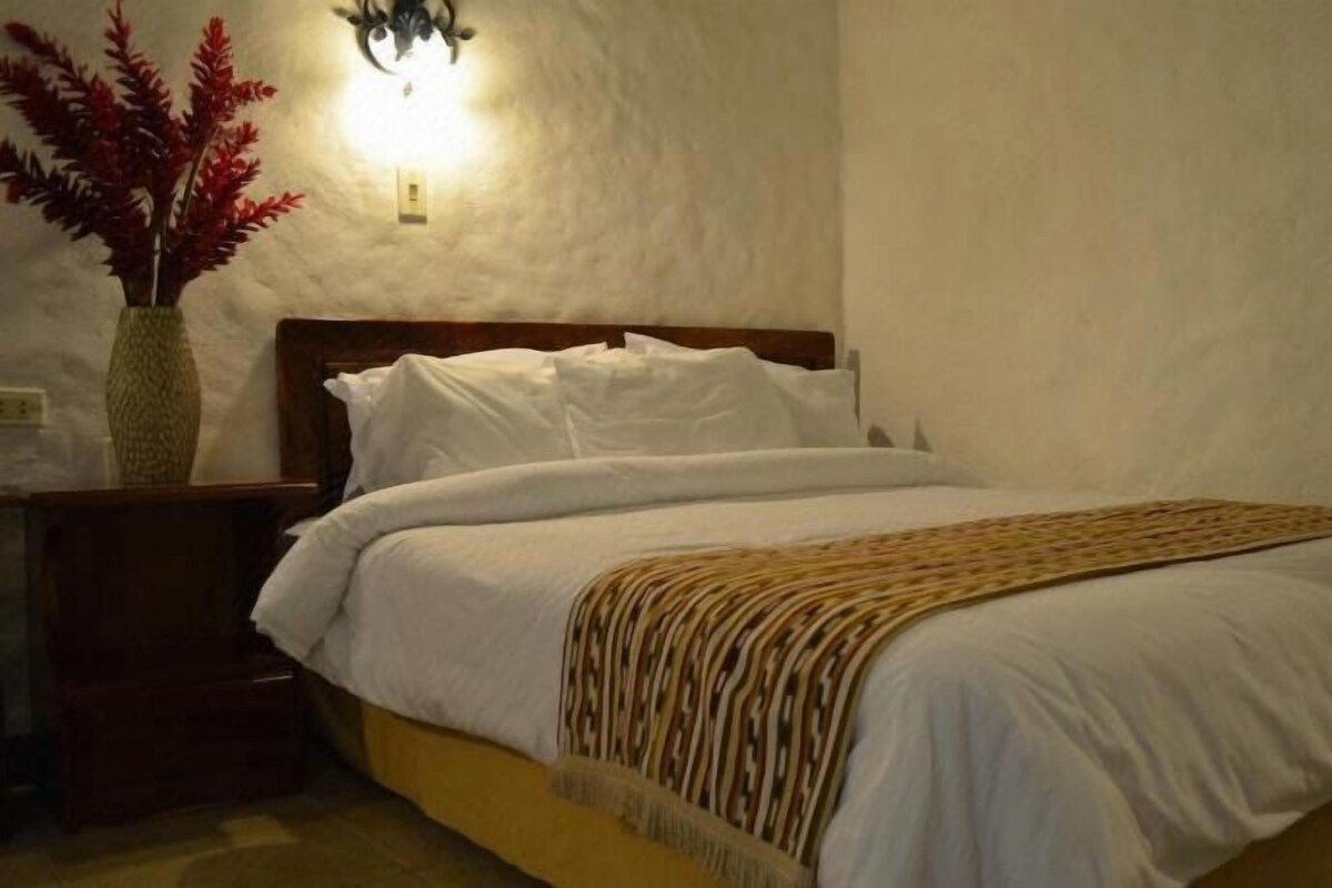 A single bed with white bedding and a patterned throw, a bedside table with a vase, and a wall-mounted light fixture.