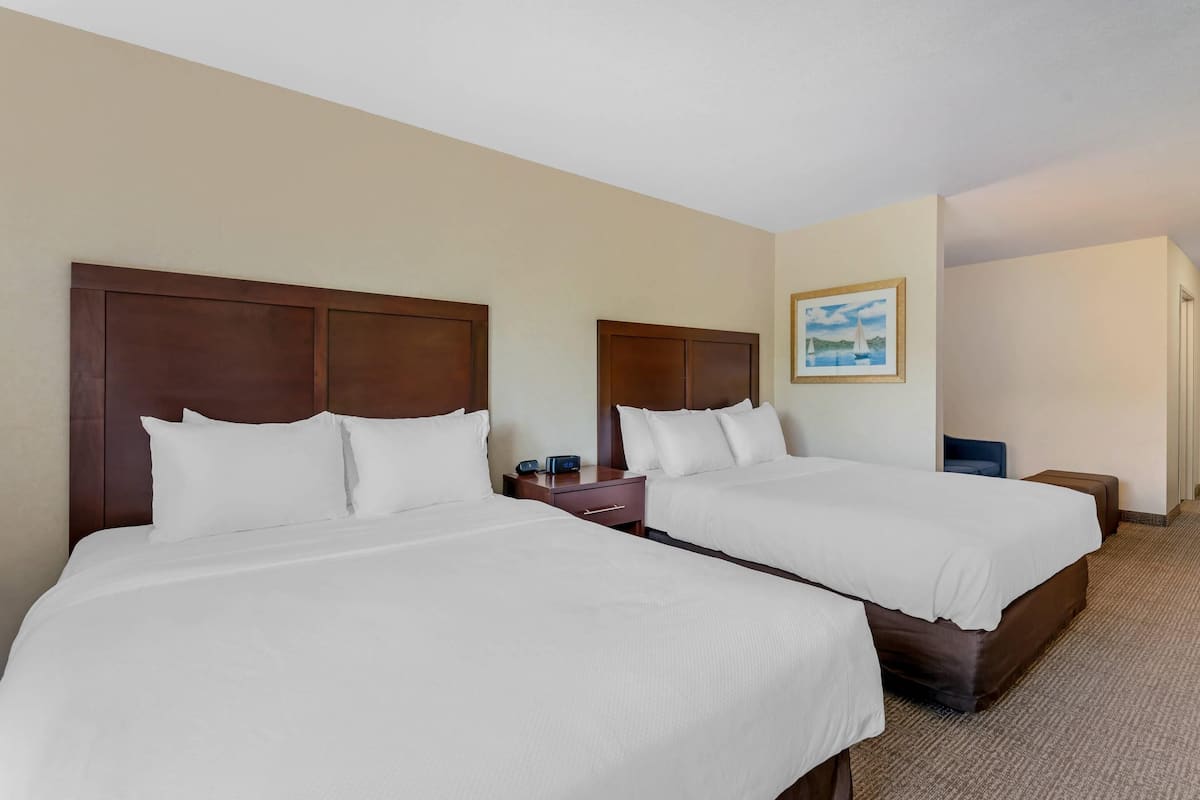 A hotel room with two beds, wooden headboards, white bedding, a nightstand with a clock, and a framed picture on the wall.
