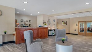 Een receptiezone met een houten balie, twee grijze fauteuils, een ronde salontafel en een klok aan de muur.