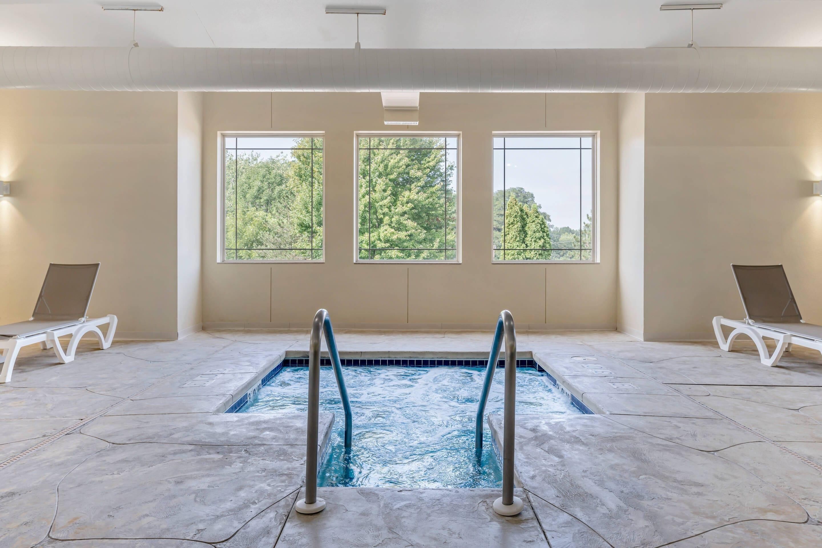 Una piscina interior con vistas despejadas a árboles a través de amplias ventanas, rodeada de sillones blancos.