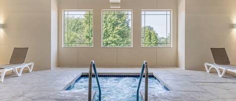 Una piscina interior con vistas despejadas a árboles a través de amplias ventanas, rodeada de sillones blancos.