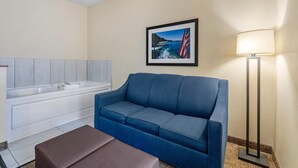 Hotelkamer met een blauwe bank, een klein bruin poufje, een witte badkuip en een lijst met een afbeelding van een brug en schip aan de muur.