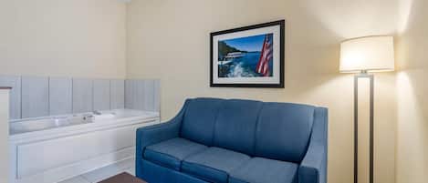 A hotel room with a blue sofa, a small brown ottoman, a white bathtub, and a framed picture of a bridge and ship on the wall.