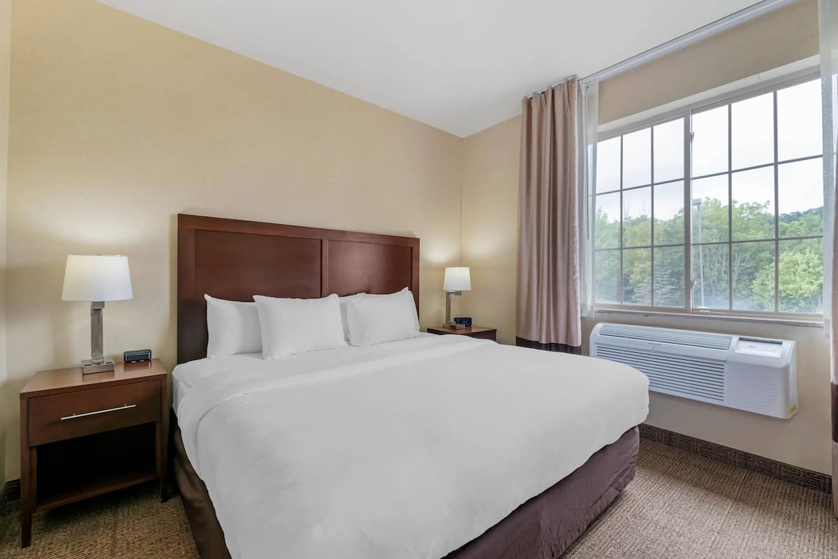 A hotel room with a large bed, two bedside tables with lamps, a window with curtains, and an air conditioning unit.