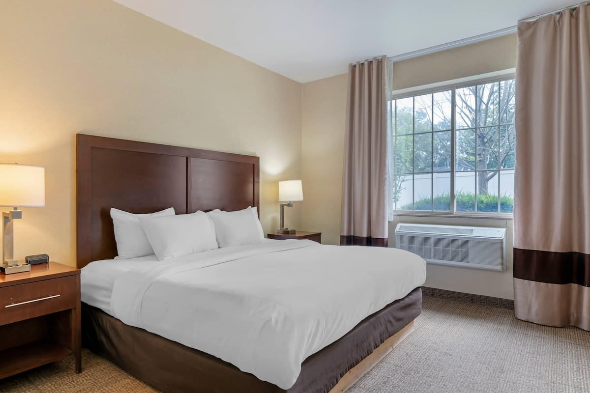 A hotel room with a large bed, two bedside tables with lamps, a window with curtains, and an air conditioning unit.