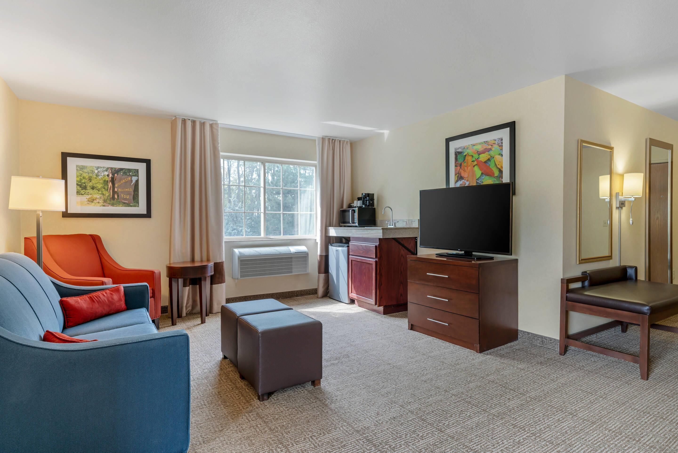 Habitación de hotel con sofá, reposapiés, una pequeña mesa, televisor, escritorio con silla y ventana con cortinas.