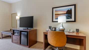 A hotel room with a desk, a television, a microwave, a refrigerator, a chair, a lamp, and a framed picture on the wall.