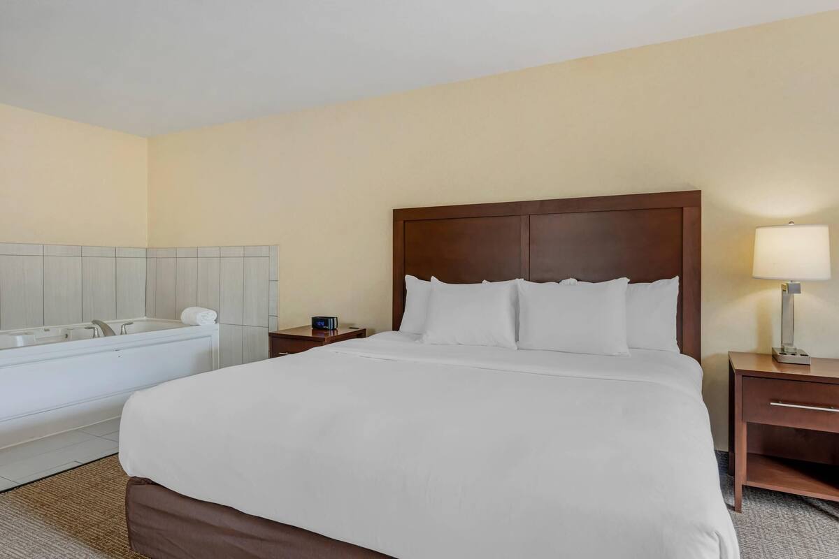 A hotel room with a large bed, a wooden headboard, a nightstand with a lamp, a small table, and a bathtub.