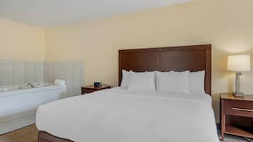 A hotel room with a large bed, a wooden headboard, a nightstand with a lamp, a small table, and a bathtub.
