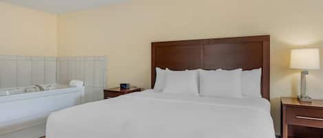 Quarto de hotel com uma cama grande, uma cabeceira de madeira, uma mesa de cabeceira com uma luminária, uma pequena mesa e uma banheira.