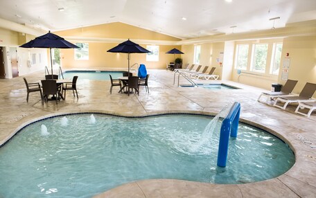 An indoor swimming pool with lounge chairs and umbrellas, surrounded by a tiled floor and walls.