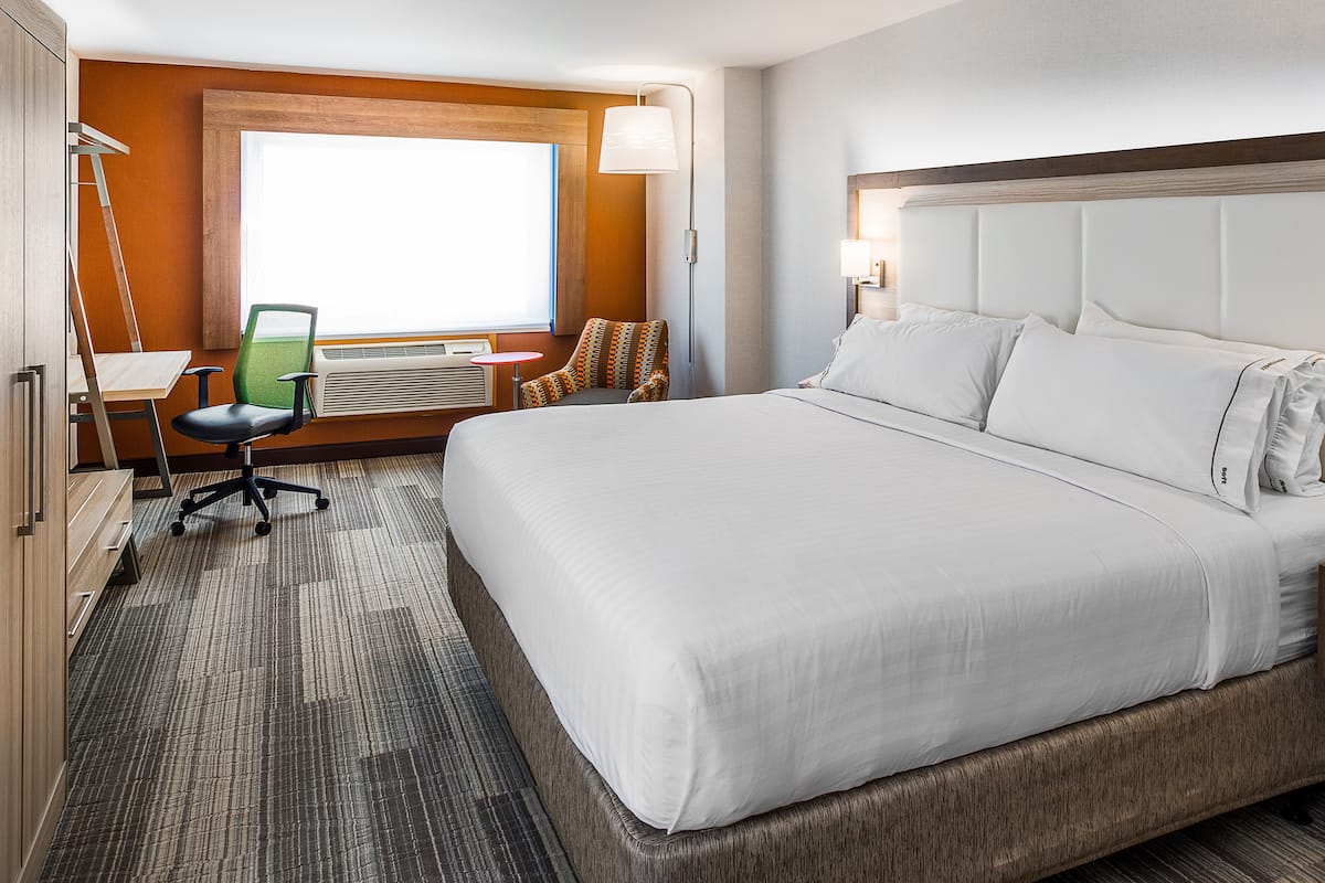 A hotel room with a large bed, a desk with a chair, an air conditioning unit, and a window with a view.