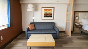 A modern hotel room with a blue sofa, a wooden coffee table, a framed artwork on the wall, and a bed with a wooden headboard. - Holiday Inn Express & Suites Halifax - Bedford by IHG (Halifax)