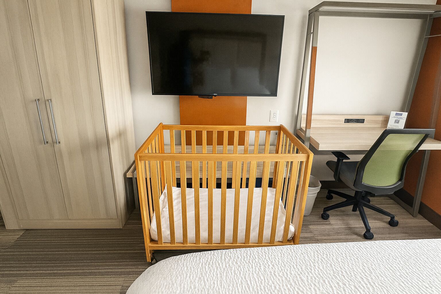 A hotel room with a wooden crib, a flat-screen TV mounted on the wall, a desk with a chair, and a bed with a white textured cover.