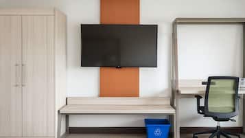A room with a TV mounted on the wall, a desk with a chair, a cabinet, and a blue recycling bin.