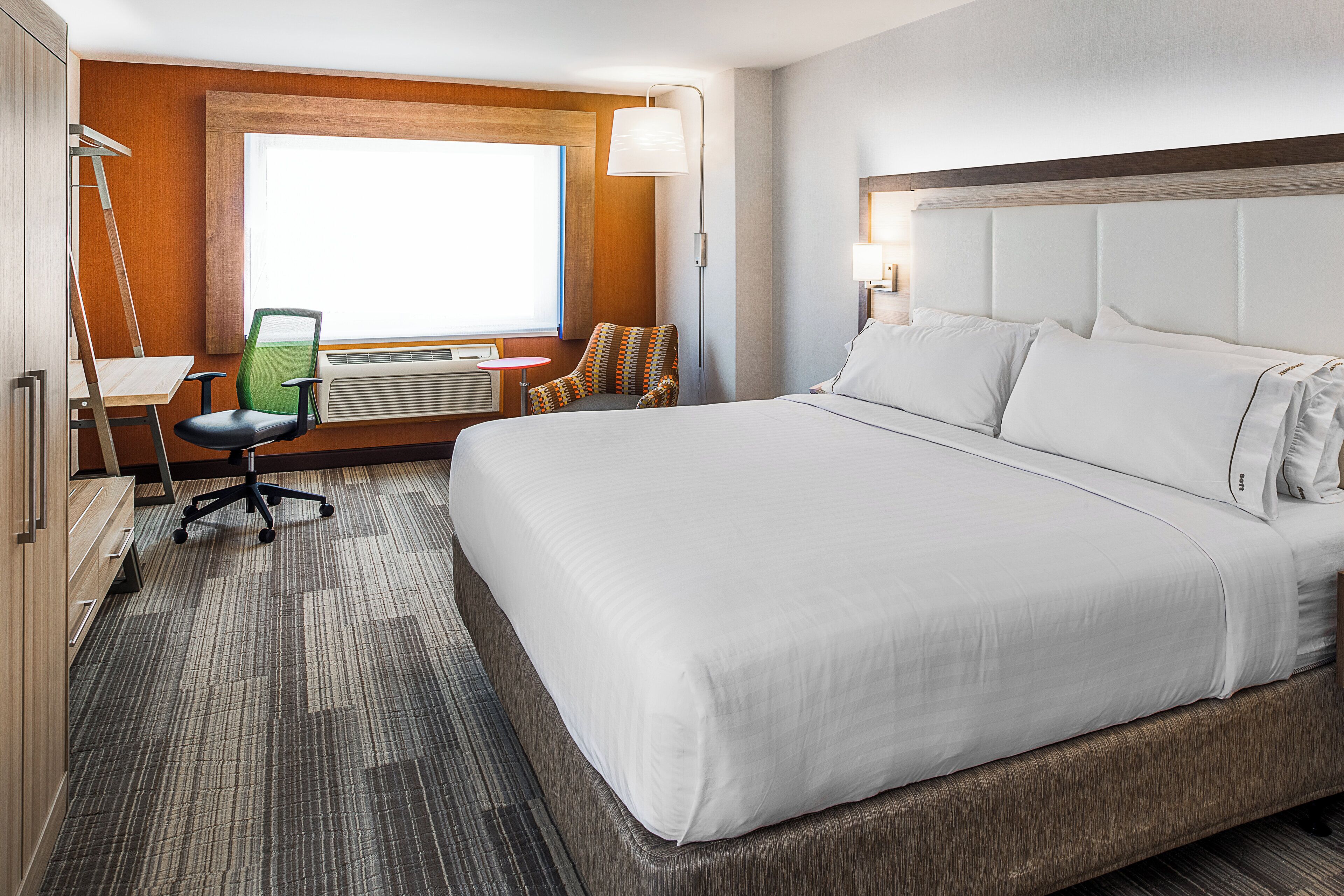 A hotel room with a large bed, a desk with a chair, a window, and a wall-mounted lamp.