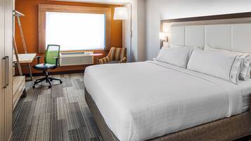 A hotel room with a large bed, a desk with a chair, a window, and a wall-mounted lamp.