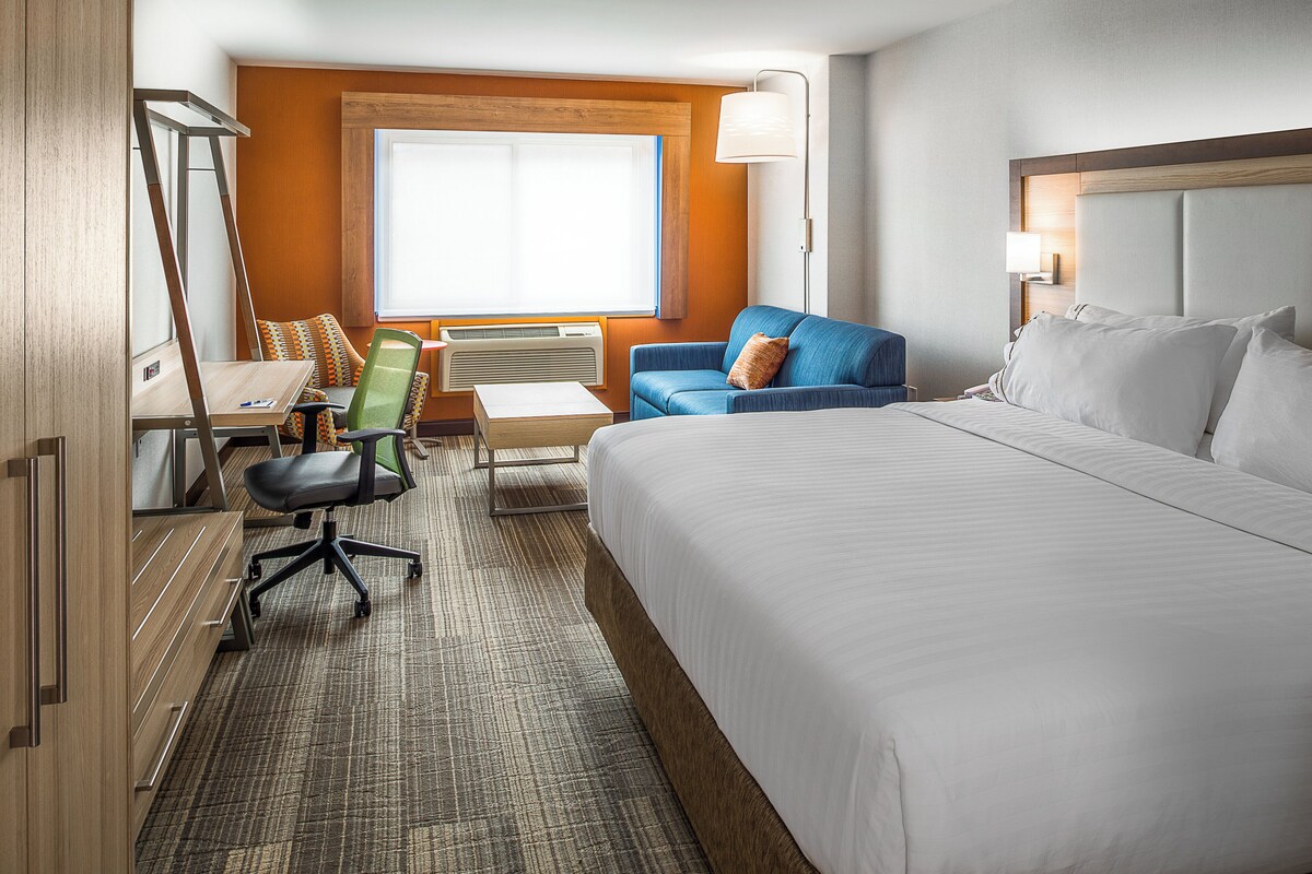A hotel room with a large bed, a desk with a chair, a sofa, and a window with blinds.