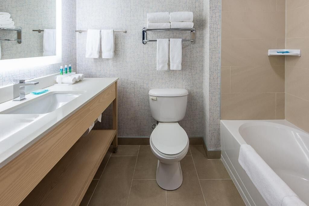 A modern bathroom with a white sink, toilet, and bathtub. There are towels hanging on a rack and a shelf with toiletries.