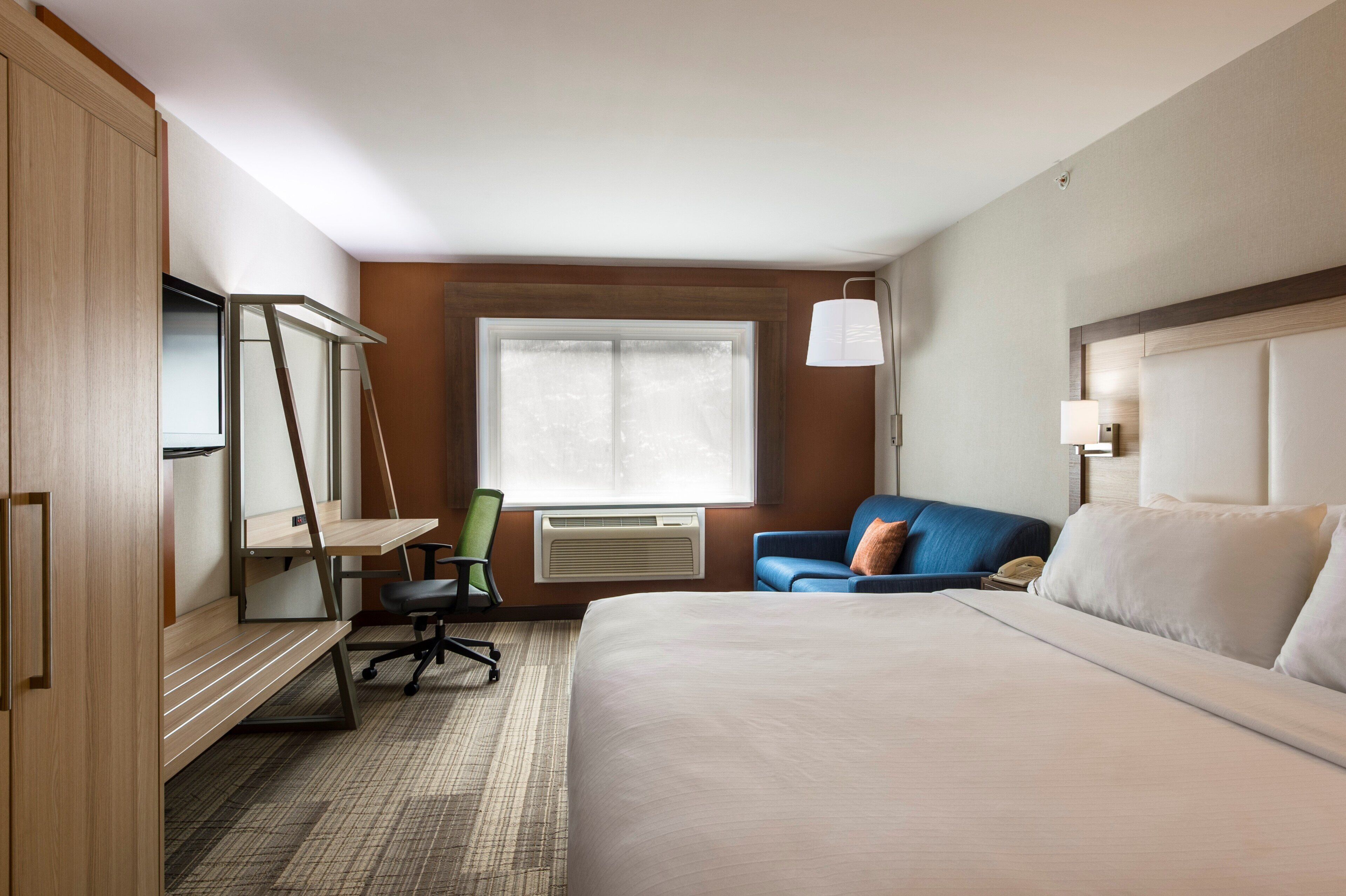 A hotel room with a bed, a desk, a chair, a window, and a lamp.