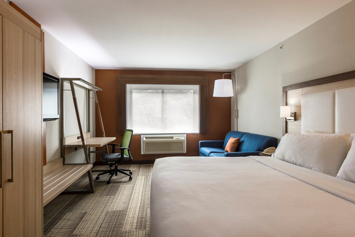 A hotel room with a bed, a desk, a chair, a window, and a lamp.