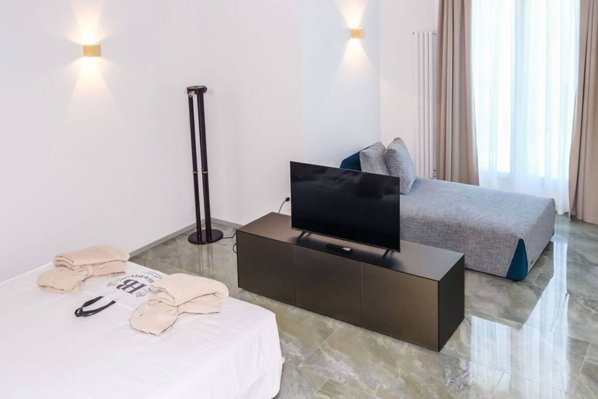 Een moderne hotelkamer met een bed, een flatscreen televisie op een donkere console, een staande lamp en een raam met gordijnen.