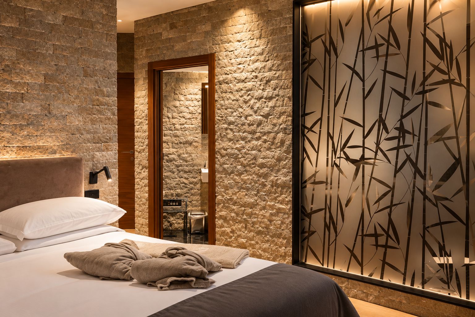 Habitación de hotel con pared de piedra, cama con almohadas y manta, mesita de noche con lámpara y separador con motivos de bambú.