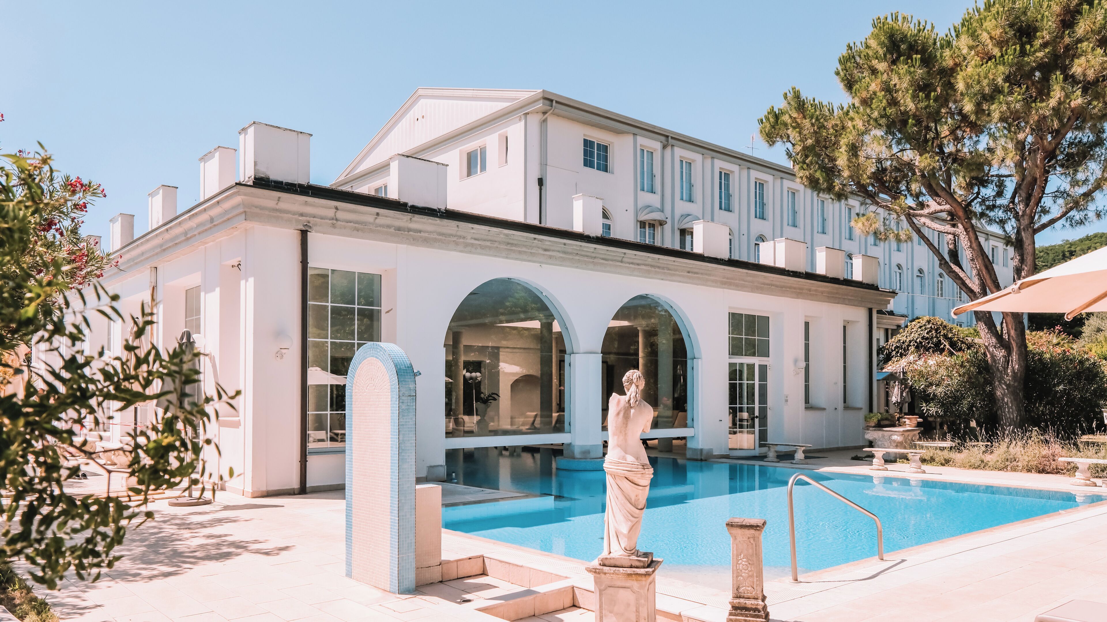 Un grand bâtiment blanc doté d’une piscine, de fenêtres en arc de cercle et d’une statue en premier plan.