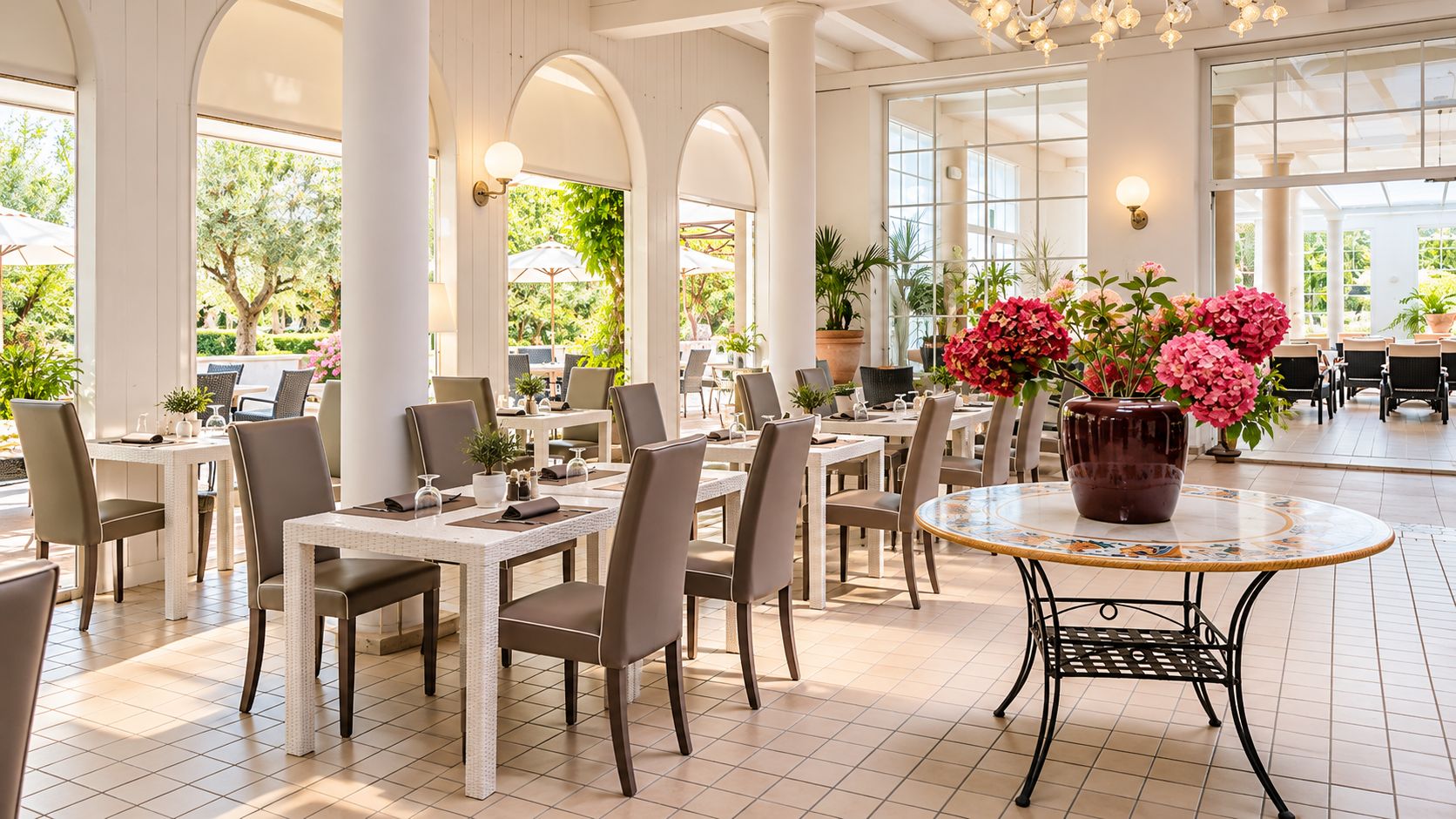 A spacious indoor dining area with multiple tables and chairs, large windows allowing natural light, and potted plants adding greenery.